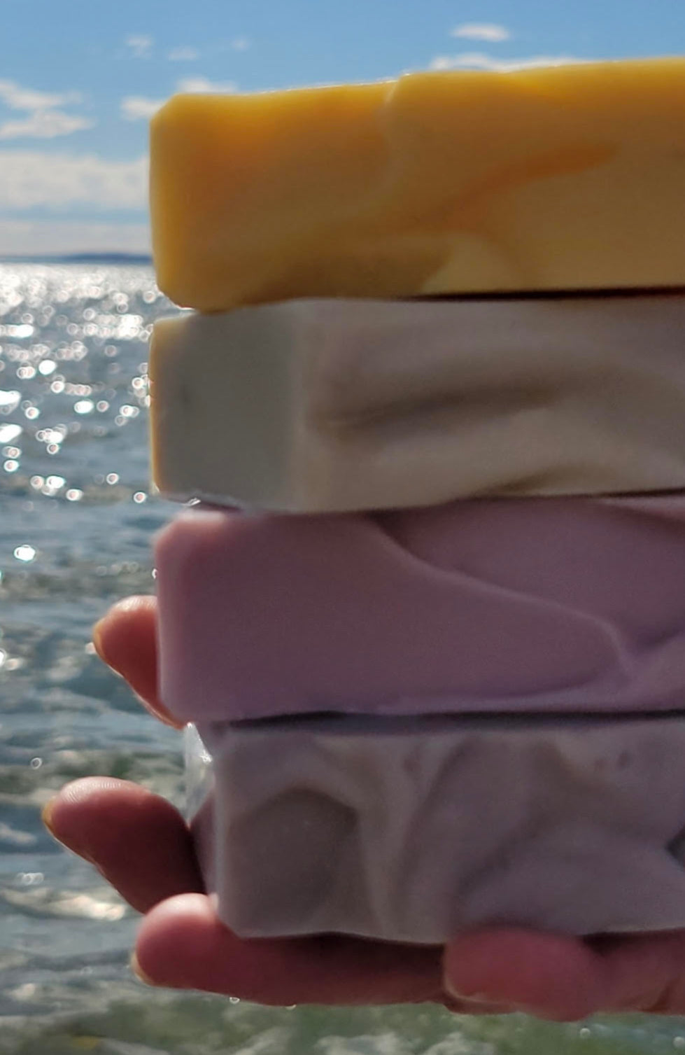 A close up view of a hand holding a stack of four soap bars with ocean waves in the background.
