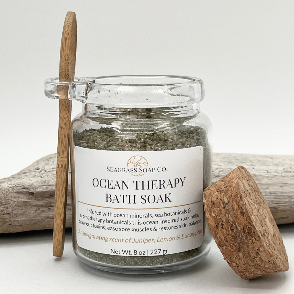 Jar of Ocean Therapy Bath Soak with a wooden spoon and cork and spoon sitting next to driftwood on a white background