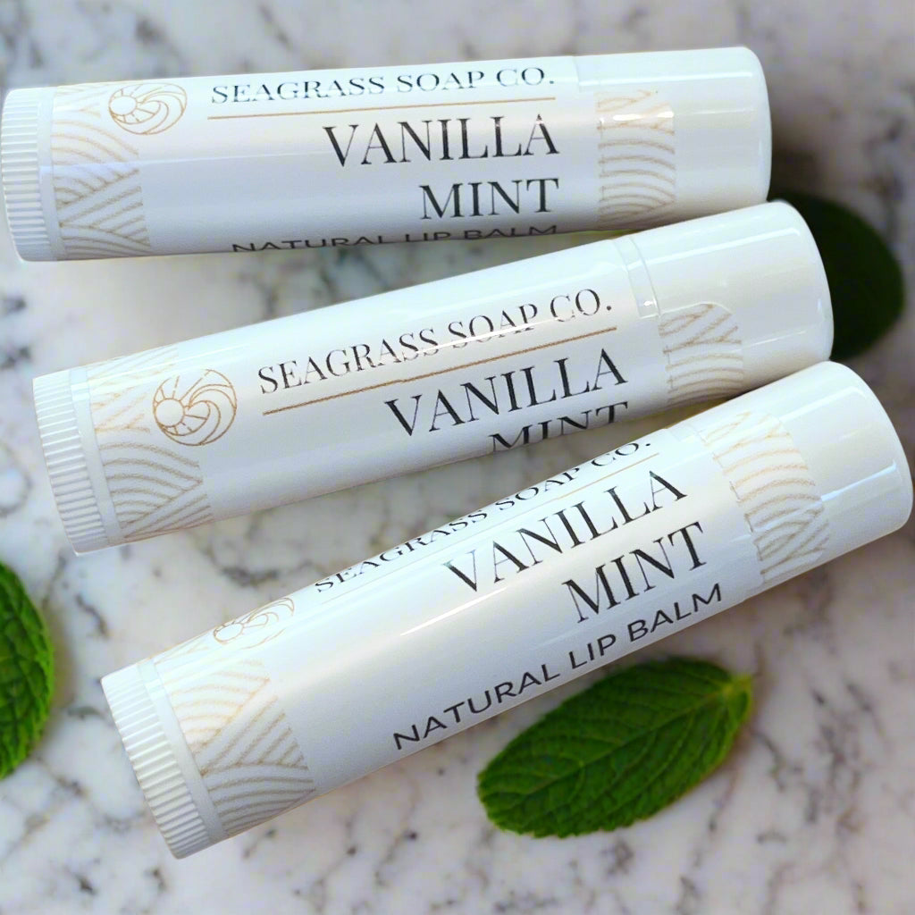 Three lip balm tubes labeled 'Vanilla Mint' from Seagrass Soap Co. on a marbeled surface with a few mint leaves surrounding them.