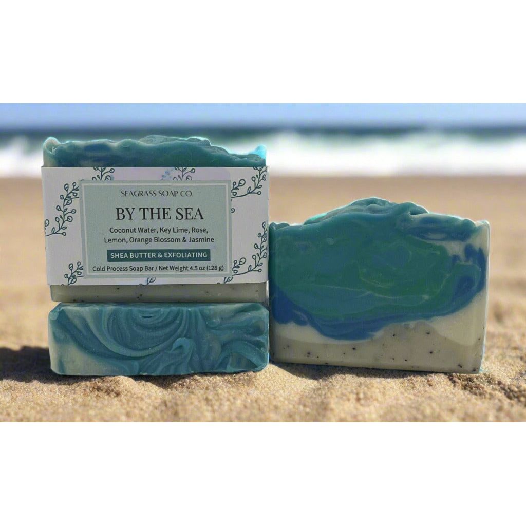 Three bars of soap resting on the sand by the waters edge. One bar is labeled and sitting on top of another showing the top design. A third bar shows the theme of a shoreline with swirls of white and blues with a small sandy beach with specks of coffee grounds which are used as a light exfoliant.