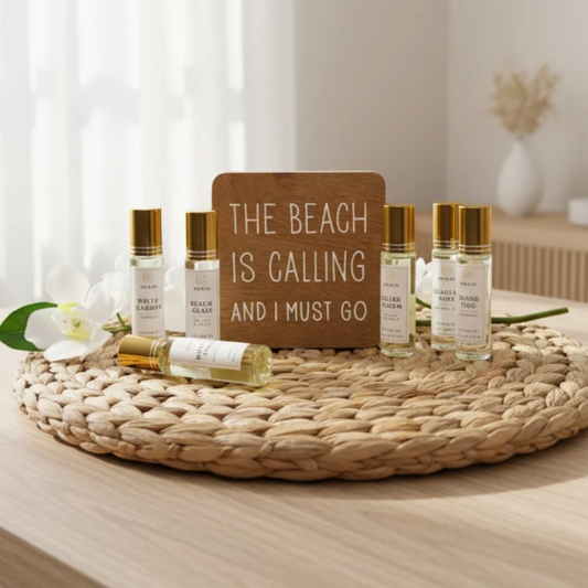 A set of five small travel perfume roller bottles with gold caps and white labels arranged on a round woven straw mat on a light wood table. A wooden sign in the center reads 'THE BEACH IS CALLING AND I MUST GO'.