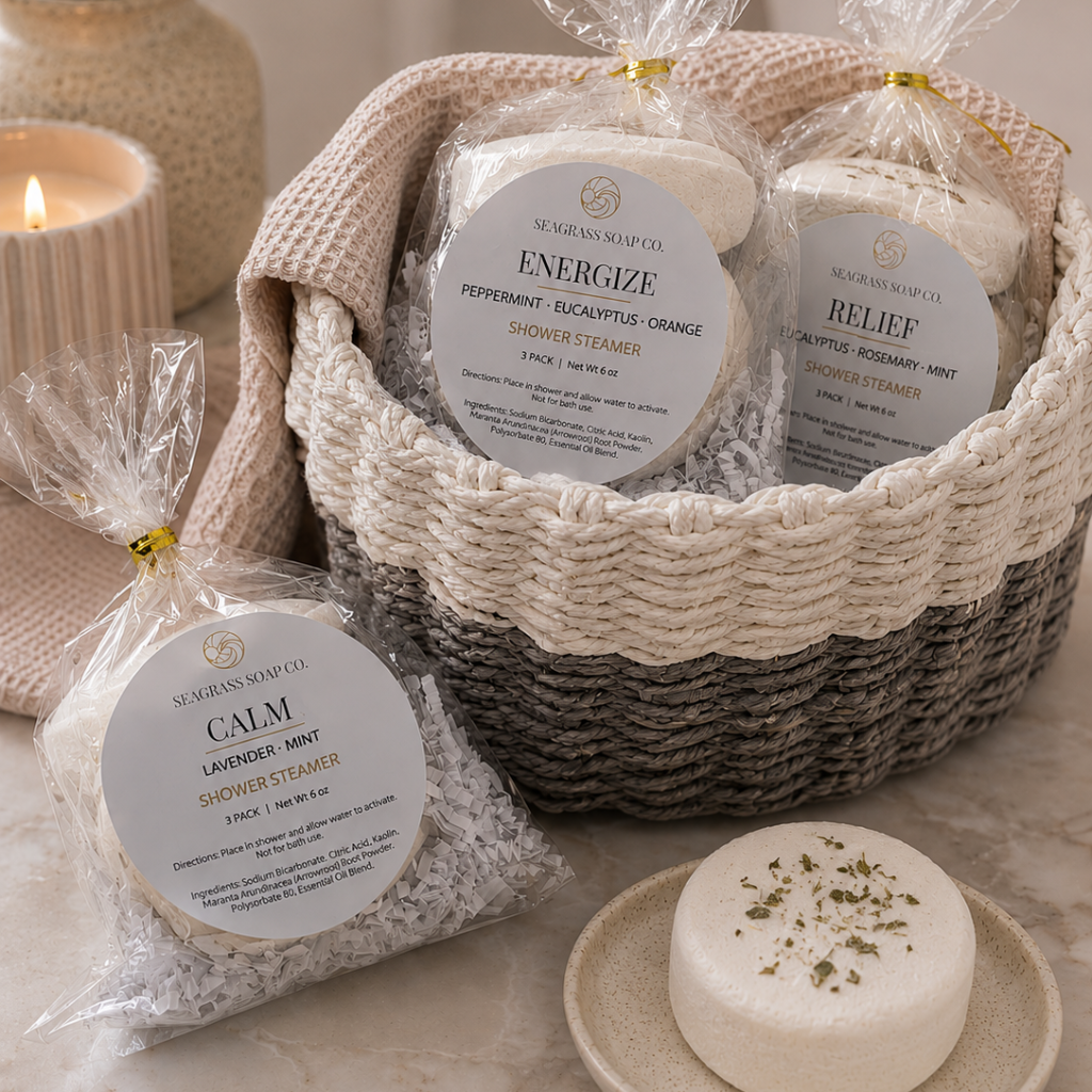 Basket of shower steamers packages displayed in a white and grey basket in a bathroom setting. One unwrapped shower steamers with botanicals sits on a round tray on a marbled counter.