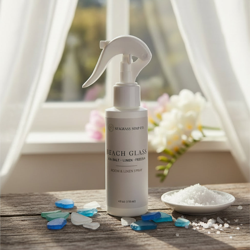 Room and linen spray on a wooden surface surrounded by florals, sea glass and sea salt in front of a window