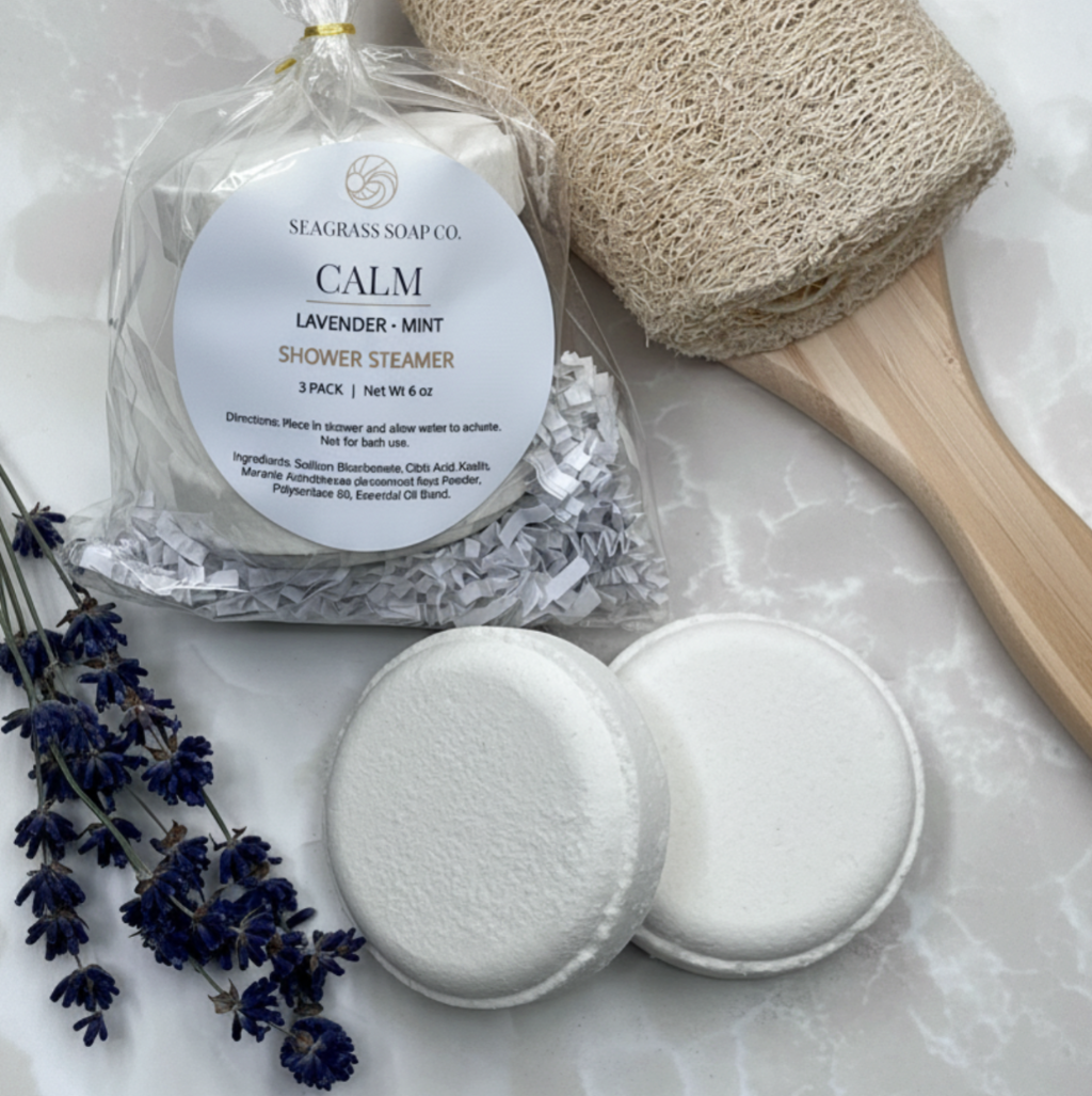 A calm shower steamer package displayed with 2 white shower steamers, a loofah brush and lavender botanicals on a marbled surface.