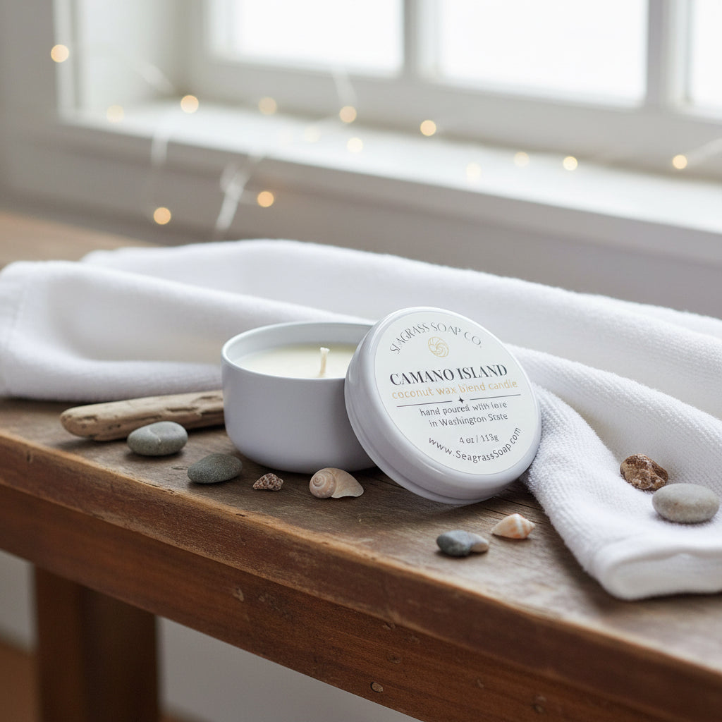 A candle tin sitting on a wooden bench with a white towel, sea shells, rocks and a piece of driftwood with a window and tiny lights in the background by Seagrass Soap Co