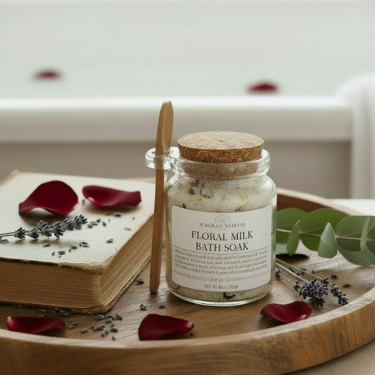 Jar of floral milk bath soak with a wooden scoop sitting on a wooden tray with an old book, rose petals and lavender stems with a bathtub in the background.