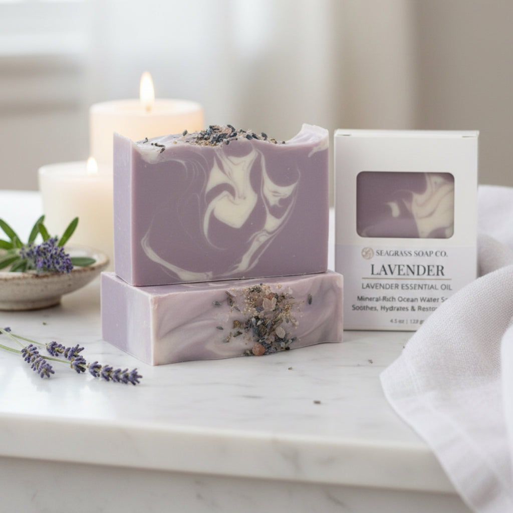 Lavender soap sitting on a marble counter next to sprigs of lavender, a towel and candles burning in the background