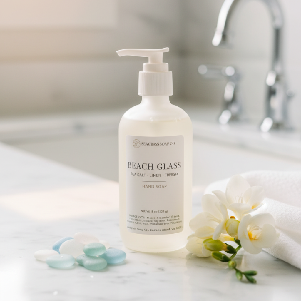 Hand soap bottle labeled 'Beach Glass' on a white surface surrounded by beach glass and florals next to a sink