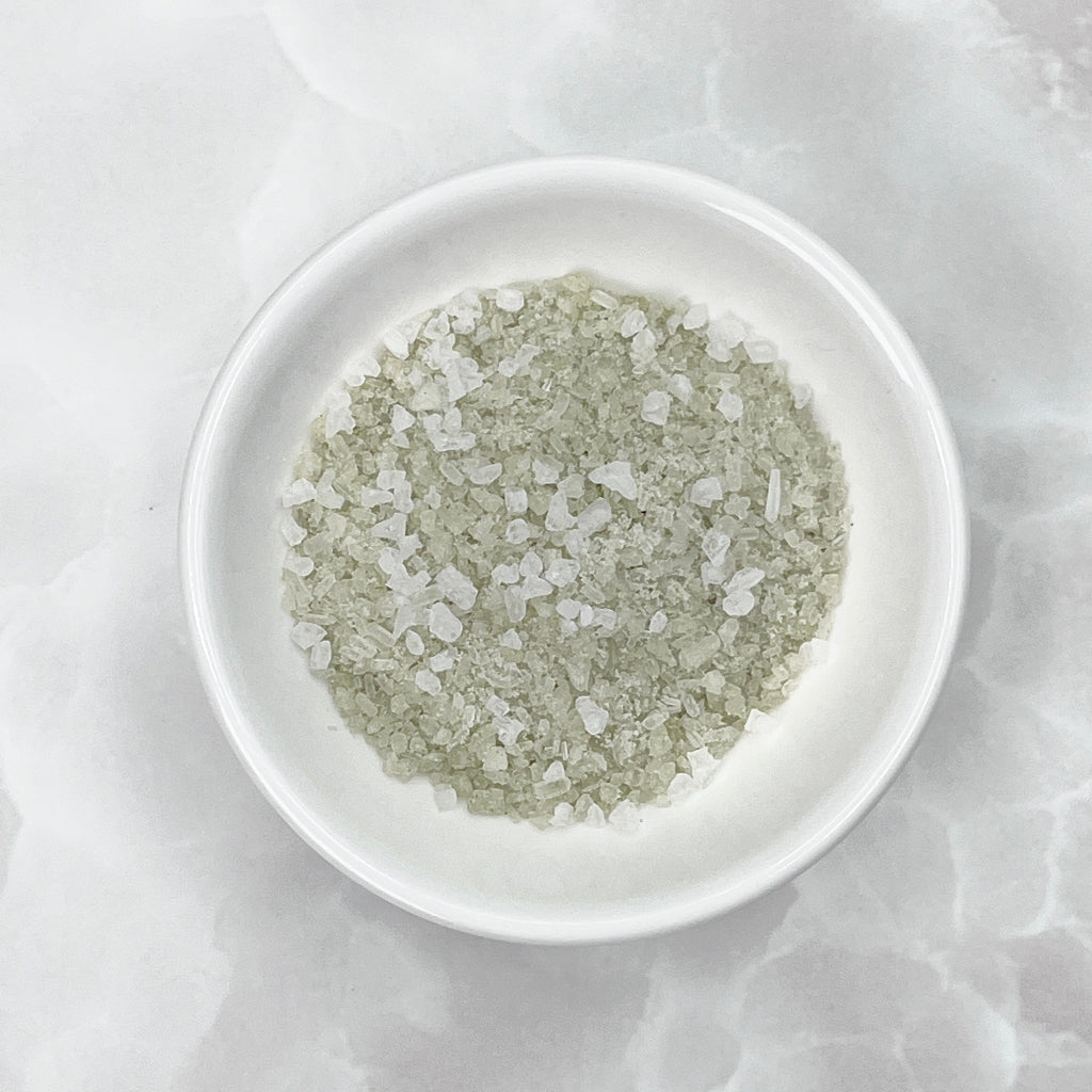 Coarse sea salt crystals with a light green tint, displayed in a small white ceramic bowl on a light gray marble surface.