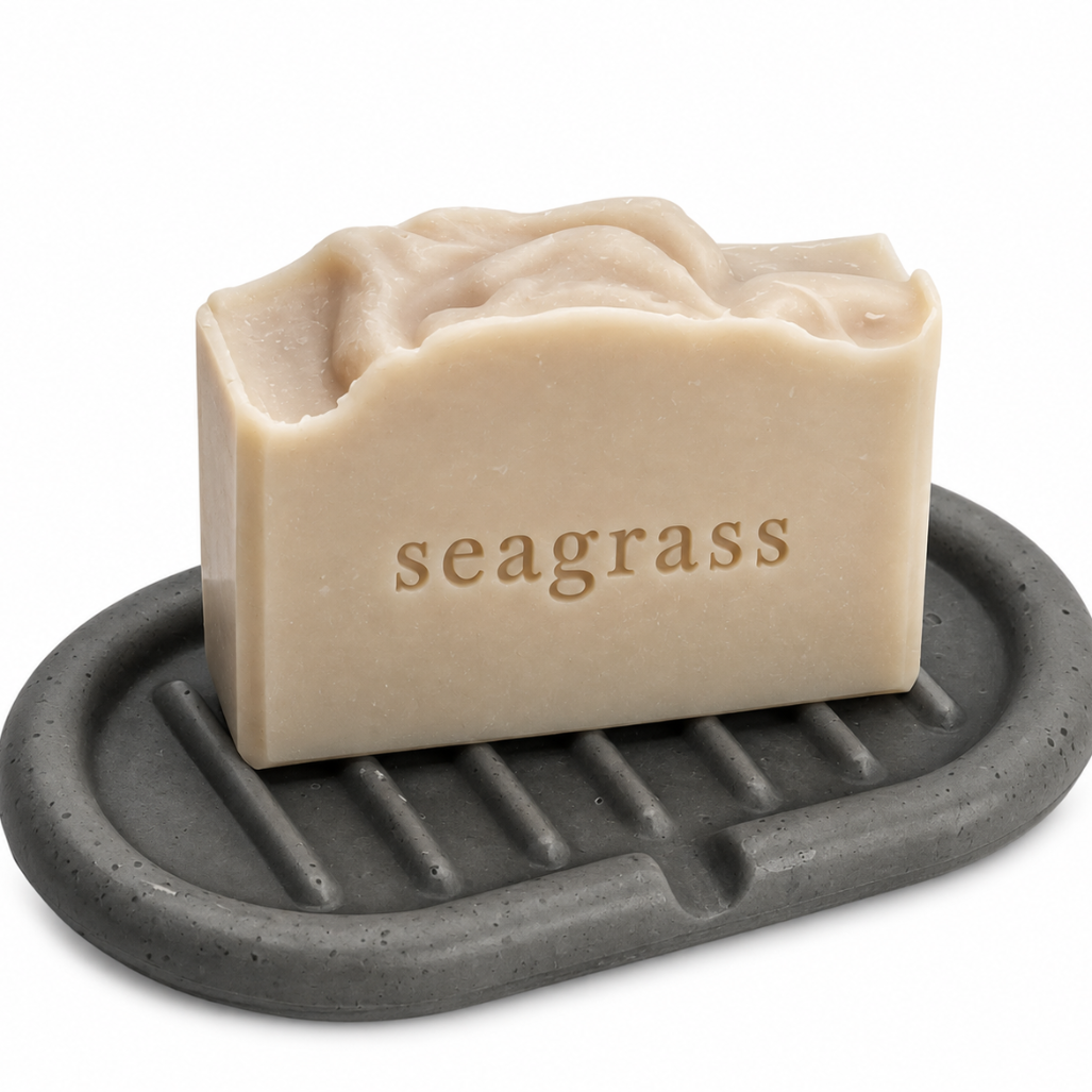 A rectangular bar of beige seagrass soap with the brand name 'seagrass' embossed on the front, resting on a dark gray oval concrete soap holder with slotted ridges, set against a plain white background.