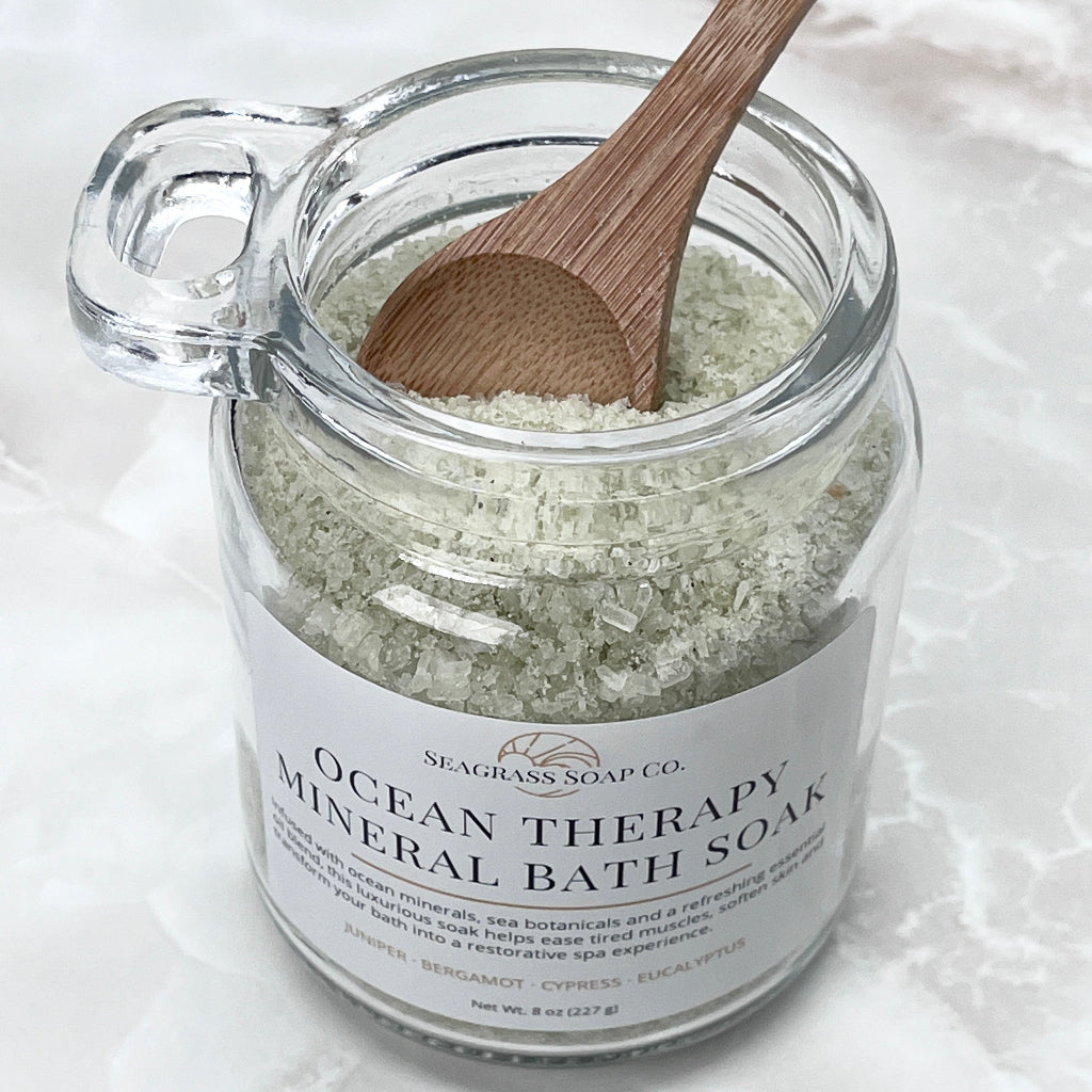 A glass jar of Seagrass Soap Co. Ocean Therapy Mineral Bath Soak with a white label and a wooden spoon resting inside, set on a light marble surface.