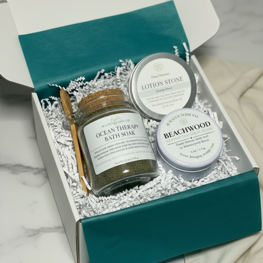 A white gift box with teal tissue containing a jar of ocean therapy bath soak, a wooden spoon, a lotion stone and a white candle tin sitting on a marble counter