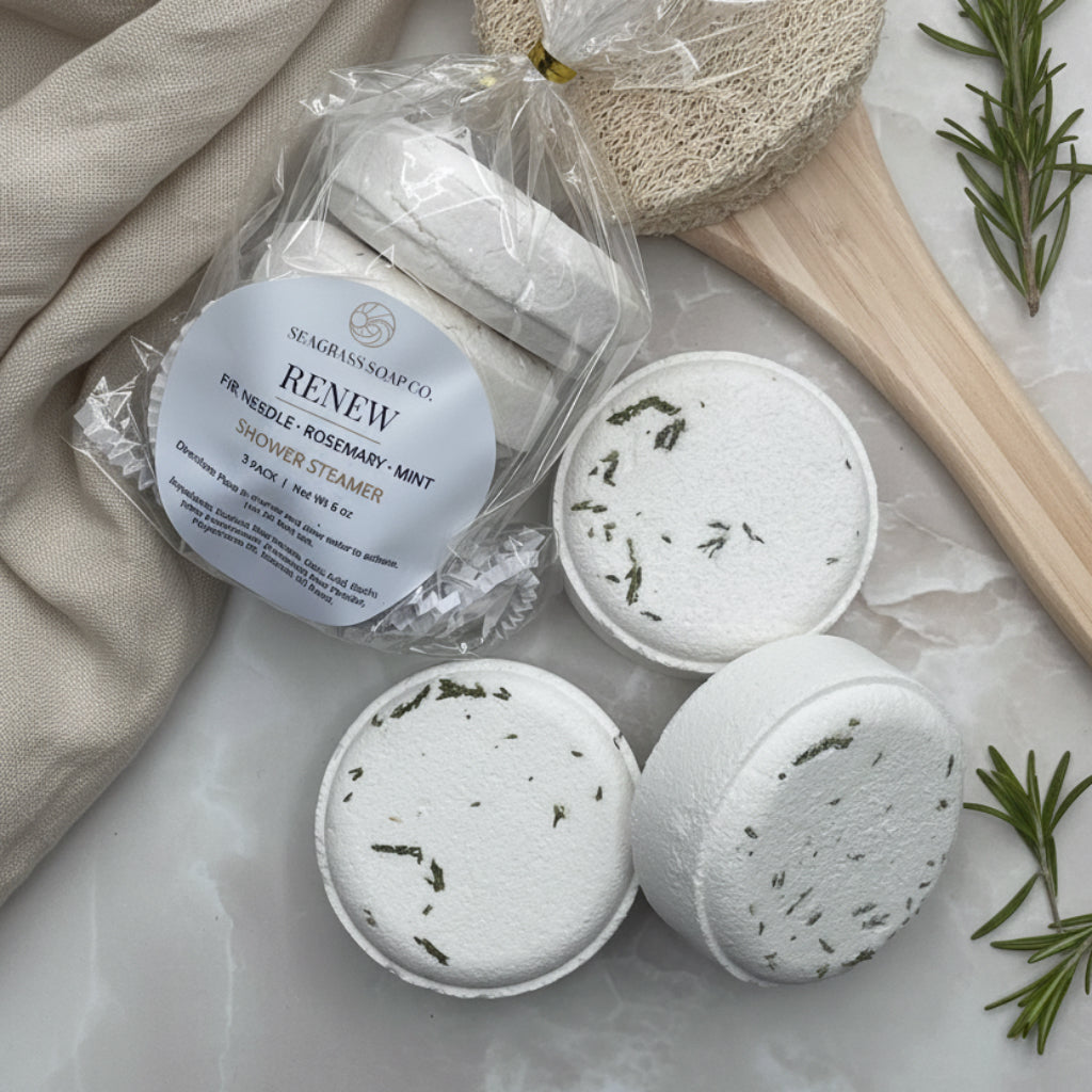 Renew shower steamer package displayed next to 3 unwrapped steamers, a towel, loofah brush and some rosemary branches.