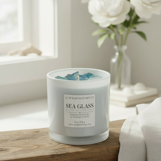 Sea Glass candle from Seagrass Soap Co. sitting on a wooden board next to a white towel with driftwood, flowers and sea shells in the background