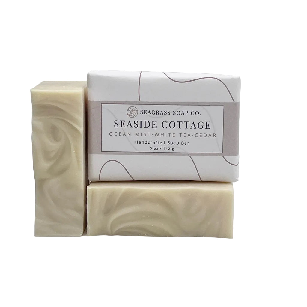Three Seaside Cottage soap bars by Seaglass Soap Co. arranged on a white background, with one wrapped bar in the center and two unwrapped cream-colored bars showing a swirled texture.